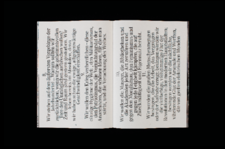 Ein offenes Buch liegt flach und zeigt zwei Seiten dichten deutschen Text, der in großen, fetten Buchstaben auf schwarzem Hintergrund gedruckt ist.