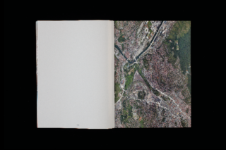 Ein offenes Buch mit einer leeren linken Seite und einem Satellitenbild einer Stadt mit einem Fluss und gitterartigen Straßen auf der rechten Seite, alles vor einem schwarzen Hintergrund.
