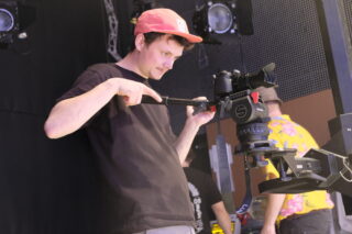 Ein Mann mit einer roten Mütze justiert eine auf einem Stativ montierte professionelle Kamera, mit Bühnenbeleuchtung und einer weiteren Person im Hintergrund.