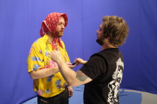 Vor einem blauen Hintergrund stehen zwei Männer. Einer trägt ein gelbes Hemd mit Blumenmuster und einen roten Schal auf dem Kopf, der andere gestikuliert mit erhobenen Händen in seine Richtung.