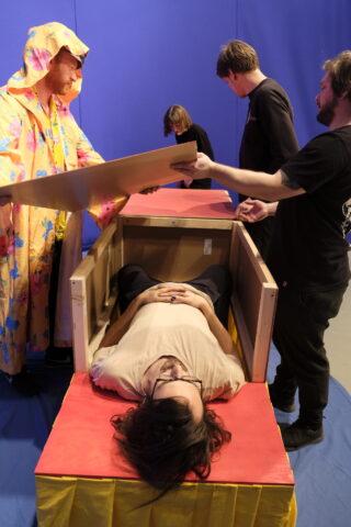 In einem Raum mit blauen Wänden liegt eine Person mit gefalteten Händen in einer Holzkiste, während drei andere, eine davon in einem bunten Gewand, den Deckel über ihr positionieren.