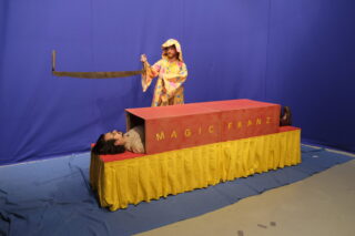 Eine Person in einem bunten Gewand hält eine große Säge über eine andere Person, die in einer Kiste mit der Aufschrift „MAGIC FRANZ“ vor blauem Hintergrund liegt.