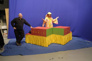 Eine Person in einem bunten Kostüm steht hinter zwei Kisten mit der Aufschrift „Magic“ und „Bravo“ auf einer Plattform mit gelbem Rock, während eine weitere Person in dunkler Kleidung neben ihnen vor einem blauen Hintergrund steht.
