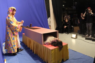 Eine Person in einem bunten Gewand steht neben einer Kiste, in der eine weitere Person liegt, während ein Kamerateam die Szene in einem Studio filmt.