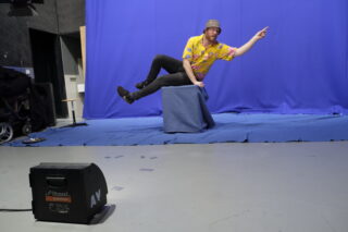 Ein Mann in buntem Hemd und Hut posiert, als würde er auf einer abgedeckten Kiste vor einem blauen Bildschirm schweben und zeigt nach vorne. Im Vordergrund ist Studioausrüstung zu sehen.