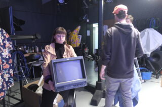 Eine Frau hält einen alten Sony-Monitor in einem mit Geräten gefüllten Studio, während zwei andere Personen in der Nähe stehen, eine mit dem Rücken zur Kamera.