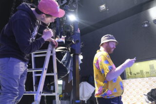 Zwei Männer arbeiten in einem Studio. Einer steht auf einer Leiter und justiert Geräte, während der andere, der ein geblümtes Hemd und einen Anglerhut trägt, auf sein Telefon schaut.