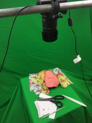 Über einem Tisch mit grünem Stoff, einem Stück Ton, gemustertem Stoff, Scheren, Papier und Bastelwerkzeugen ist vor einem Greenscreen-Hintergrund eine Kamera angebracht.