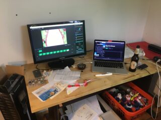 Ein überfüllter Schreibtisch mit einem Monitor, auf dem Animationssoftware angezeigt wird, einem offenen Laptop, einer Bierflasche, verstreuten Papieren, Elektronik und verschiedenen Vorräten in einer orangefarbenen Kiste darunter.