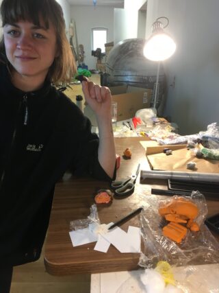 Eine Frau sitzt in einem Arbeitszimmer an einem überfüllten Tisch mit Bildhauermaterialien, Werkzeugen und Tonfiguren unter einer Schreibtischlampe.