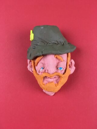 Eine Ton-Skulptur eines Männerkopfes mit grünem Hut, orangefarbenem Bart und blauen Augen vor einem einfarbig roten Hintergrund.