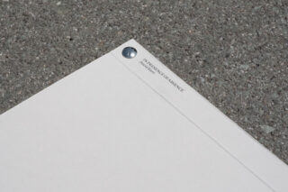 Ein weißes Dokument mit einem Metallverschluss auf einer gesprenkelten grauen Oberfläche, in der oberen Ecke mit „IN PRESENCE OF ABSENCE“ betitelt.