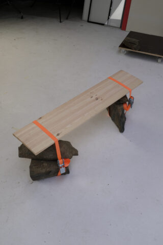 Ein von Felsen gestütztes und mit orangefarbenen Bändern befestigtes Holzbrett bildet eine provisorische Bank auf einem hellen Boden.