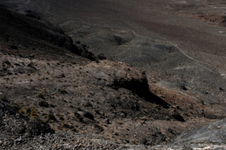 Ein felsiger, karger Hang mit spärlicher Vegetation erstreckt sich bei bedecktem Himmel in eine trockene, karge Landschaft.