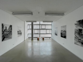 Ein minimalistischer Galerieraum mit großen Fenstern, weißen Wänden, ausgestellten Schwarzweiß-Landschaftsfotografien und zwei orangefarbenen Hockern in der Nähe des mittleren Fensters.