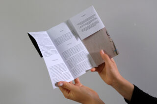 Eine Person hält und liest eine gefaltete Broschüre mit gedrucktem Text und einem Teilbild auf weißem Hintergrund.