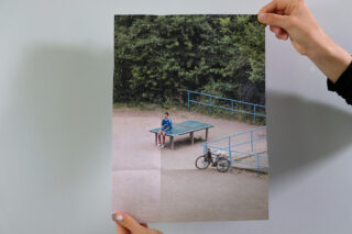 Eine Person hält ein Foto von einem Mann, der allein an einer Tischtennisplatte im Freien sitzt. In der Nähe steht ein Fahrrad und im Hintergrund sind Bäume zu sehen.