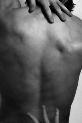 Schwarzweißfoto, das den nackten Rücken einer Person zeigt, auf deren Schultern und unterem Rücken die Hände einer anderen Person ruhen.