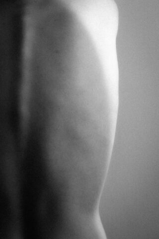 Schwarzweißfoto des nackten Rückens einer Person mit sichtbarer Wirbelsäule und Schulterblatt, schwach beleuchtet und unscharf.