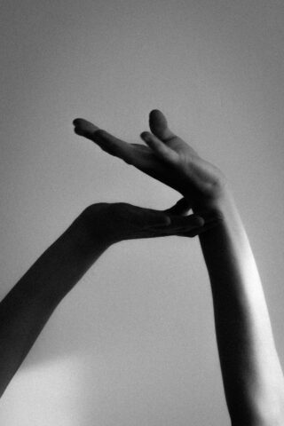 Schwarzweißfoto von zwei menschlichen Armen und Händen, die vor einem schlichten Hintergrund erhoben sind, die Finger anmutig gekreuzt und ausgestreckt.
