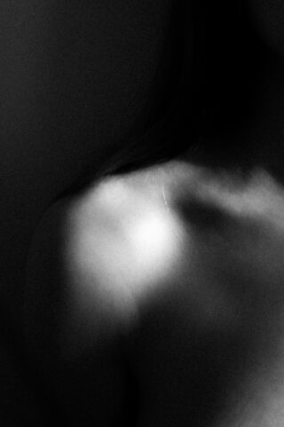 Schwarzweißfoto, das die nackte Schulter und das Schlüsselbein einer Person zeigt, die von sanftem Licht beleuchtet werden, wobei die Schatten einen dramatischen Kontrast erzeugen.