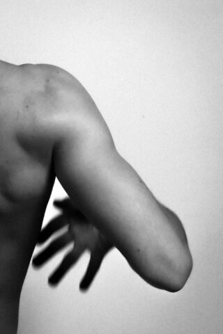 Schwarzweißfoto, das den oberen Rücken, die Schulter und den Arm einer Person vor einem einfarbigen Hintergrund zeigt, wobei der Arm angewinkelt und die Hand in Bewegung leicht unscharf ist.
