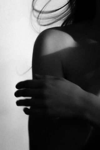 Schwarzweißbild, das eine Nahaufnahme der Schulter und Hand einer Person mit teilweise sichtbarem Haar zeigt, beleuchtet durch weiches Licht und Schatten.