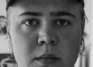 Eine Nahaufnahme in Schwarzweiß, auf der das Gesicht einer Person zu sehen ist, die eine Mütze trägt und direkt in die Kamera blickt.