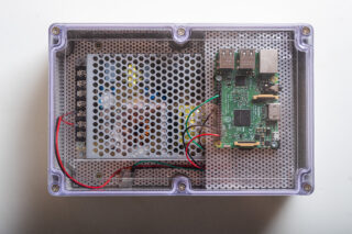 Ein kleines Elektronikgehäuse mit einem Netzteil auf der linken Seite und einem Raspberry Pi-Einplatinencomputer auf der rechten Seite, verbunden durch rote, grüne und gelbe Kabel.