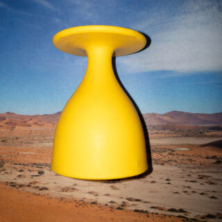 Ein gelber, umgedrehter Plastikbecher steht vor dem Hintergrund einer Wüstenlandschaft mit blauem Himmel und Bergen in der Ferne.