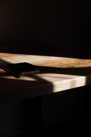 Ein geschlossenes Notizbuch und ein Stift liegen auf einem Holztisch, teilweise beleuchtet durch Sonnenlicht und sich kreuzende Schatten.