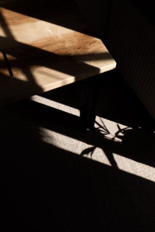 Ein Holztisch mit schwarzen Beinen wird teilweise vom Sonnenlicht beleuchtet und wirft scharfe Schatten und Pflanzensilhouetten auf einen Teppichboden.