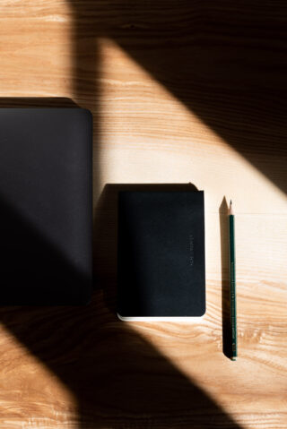 Ein geschlossener schwarzer Laptop, ein kleines schwarzes Notizbuch und ein grüner Bleistift sind auf einer Holzoberfläche angeordnet, auf der das Sonnenlicht eckige Schatten wirft.