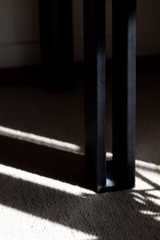 Nahaufnahme eines schwarzen Tischbeins aus Metall, das einen Schatten auf einen beigen Teppich wirft, in den von links Sonnenlicht einfällt.
