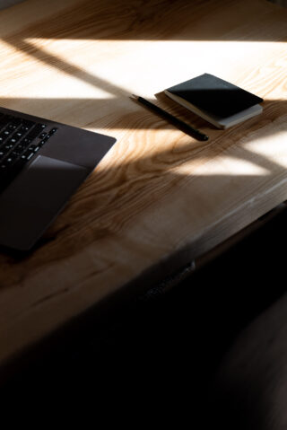 Ein Holzschreibtisch mit einem teilweise sichtbaren Laptop, einem geschlossenen schwarzen Notizbuch und einem Stift, auf dessen Oberfläche das Sonnenlicht Schatten wirft.