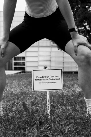 Eine Person in Shorts hockt auf einer Rasenfläche über einem Schild mit deutscher Aufschrift; das Schild verweist als feministisches Statement auf Menstruationsblut.