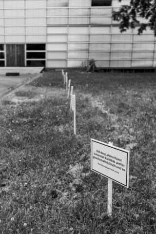 Schwarzweißfoto von Schildern mit deutschem Text, die auf einer Rasenfläche neben einem modernen Gebäude mit horizontalen Paneelen angebracht sind. Das nächstgelegene Schild ist deutlich zu sehen.