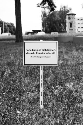 Auf einer Wiese steht auf einem Schild auf Deutsch: „Papa kann es sich leisten, dass du Kunst studierst? Mehr Klischee geht nicht, sorry.“ Im Hintergrund sind Bäume und Gebäude zu sehen.