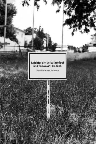 Auf einer Rasenfläche, im Hintergrund Häuser und Bäume, ist ein Schild in deutscher Sprache aufgestellt. Das Schild stellt die Verwendung ironischer und provokativer Schilder in Frage.
