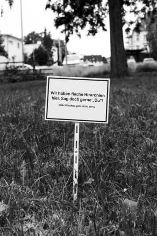 Auf einer Rasenfläche, im Hintergrund umgeben von Bäumen und Gebäuden, ist ein kleines Schild in deutscher Sprache angebracht.