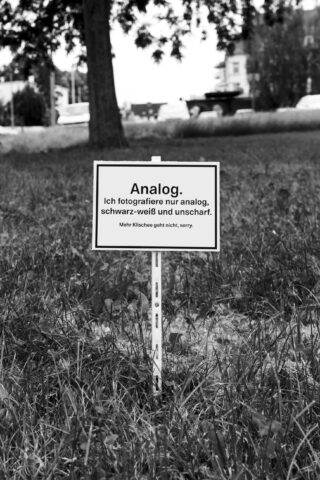 Auf einer Wiese steht ein Schild mit deutscher Aufschrift: „Analog. Ich fotografiere ausschließlich analog, schwarz-weiß und unscharf. Klischeehafter geht es leider nicht.“ Im Hintergrund sind Bäume und Gebäude zu sehen.
