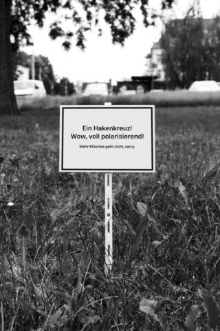 Auf einem Schild auf einer Wiese steht auf Deutsch: „Ein Hakenkreuz! Wow, voll polarisierend! Mehr Klischee geht nicht, sorry.“ Im Hintergrund sind Gebäude und Autos zu sehen.