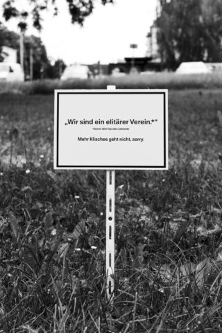 Im hohen Gras steht ein Schild auf Deutsch mit der Aufschrift: „Wir sind ein elitärer Verein. Mehr Klischee geht nicht, sorry.“ Im unscharfen Hintergrund sind Autos und Bäume zu erkennen.
