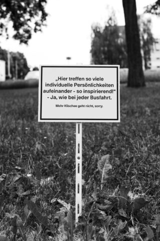 Auf einer Rasenfläche steht ein Schild in deutscher Sprache, das eine Botschaft über individuelle Persönlichkeiten und Inspirationen enthält, während im Hintergrund Gebäude und Bäume unscharf zu erkennen sind.