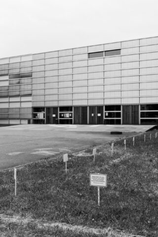 Ein modernes Gebäude mit Glasfront steht hinter einer Rasenfläche, die mit kleinen Schildern und Pfählen markiert ist. Im Vordergrund ist ein größeres Schild zu sehen. Die Szene wirkt leer und still.