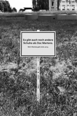 Auf einer Wiese steht ein Schild in deutscher Sprache mit der Aufschrift: „Es gibt auch noch andere Schuhe als Doc Martens. Mehr Klischee geht nicht, sorry.“ Im Hintergrund sind Gebäude zu erkennen.