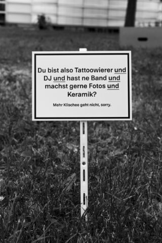 Auf einem auf dem Rasen angebrachten Schild in deutscher Sprache steht: „Du bist auch Tattoowierer und DJ und hast ne Band und machst gerne Fotos und Keramik? Mehr Klischee geht nicht, sorry.“.