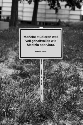 Auf einem Schild auf Deutsch steht: „Manche studieren was voll Gehaltvolles wie Medizin oder Jura. Wir halt Kunst“, aufgestellt auf einer Rasenfläche im Freien mit einem Gebäude im Hintergrund.