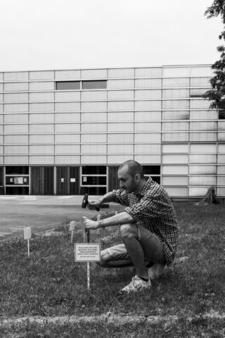 Ein Mann hockt im Gras und hämmert vor einem modernen Gebäude mit großen Fenstern ein Schild in den Boden.