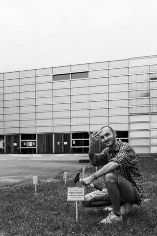 Ein Mann hockt im Gras neben einem kleinen Schild mit Text vor einem modernen Gebäude mit Glasfassade. Er gestikuliert mit einer Hand und blickt in die Kamera.